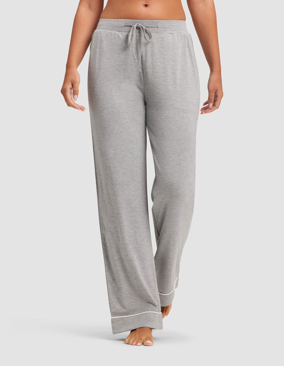 A woman wears Cozy Earth Women's Bamboo Stretch-Knit Pajama Pants in light gray, featuring a drawstring waist and white trim at the cuffs, standing barefoot against a plain background.