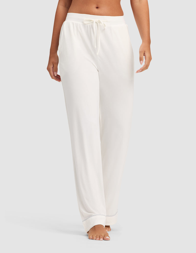 A model wears HIDE Women's Bamboo Stretch-Knit Pajama Pants in white, featuring a loose fit and drawstring waist, barefoot against a plain light gray background. Only the lower torso and legs are shown.