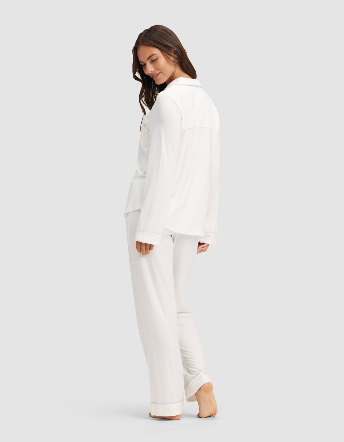 A woman with long brown hair stands barefoot, smiling and turning slightly to the side. She wears the Cozy Earth Women's Bamboo Stretch-Knit Long Sleeve Pajama Set in white, with pants, against a plain light gray background. |Color:Ivory