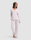 A woman smiles while wearing the Cozy Earth Women's Bamboo Stretch-Knit Long Sleeve Pajama Set, featuring a button-up top and matching pants in light lavender, standing barefoot against a plain light background. |Color:Lilac