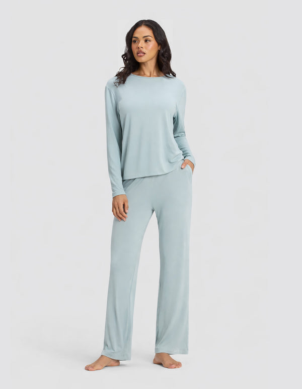 A woman stands barefoot against a plain background, wearing the Cozy Earth Women's Bamboo Stretch-Knit Long Sleeve Lounge Tee & Pant Set in Brook, with one hand in her pocket and a relaxed expression.|Color:Brook