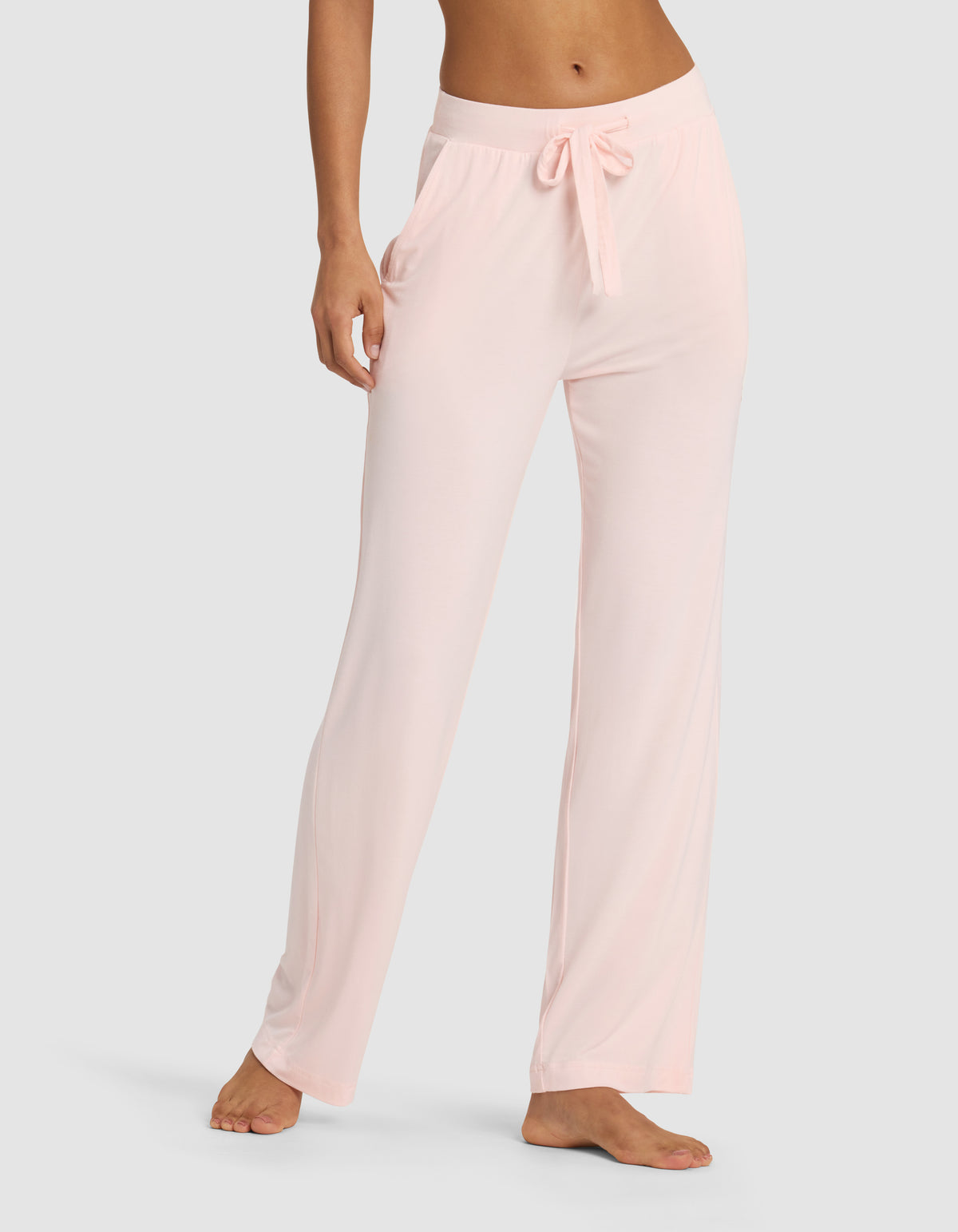 A woman wears Cozy Earth Women's Bamboo Stretch-Knit Pant in light pink, featuring a loose fit, elastic waistband, and drawstring, standing barefoot against a plain background.|Color:Blossom