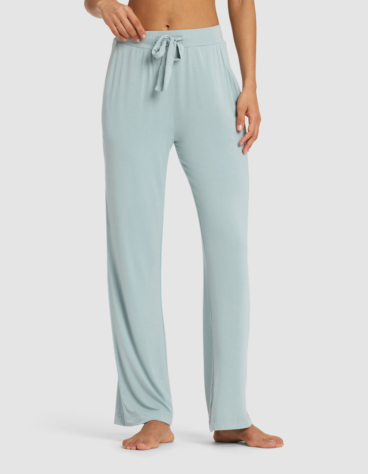 A woman models Cozy Earth’s Women's Bamboo Stretch-Knit Pant, featuring a relaxed fit, drawstring waist, and light blue color. She stands barefoot on a plain backdrop, the floor-length pants draping comfortably.