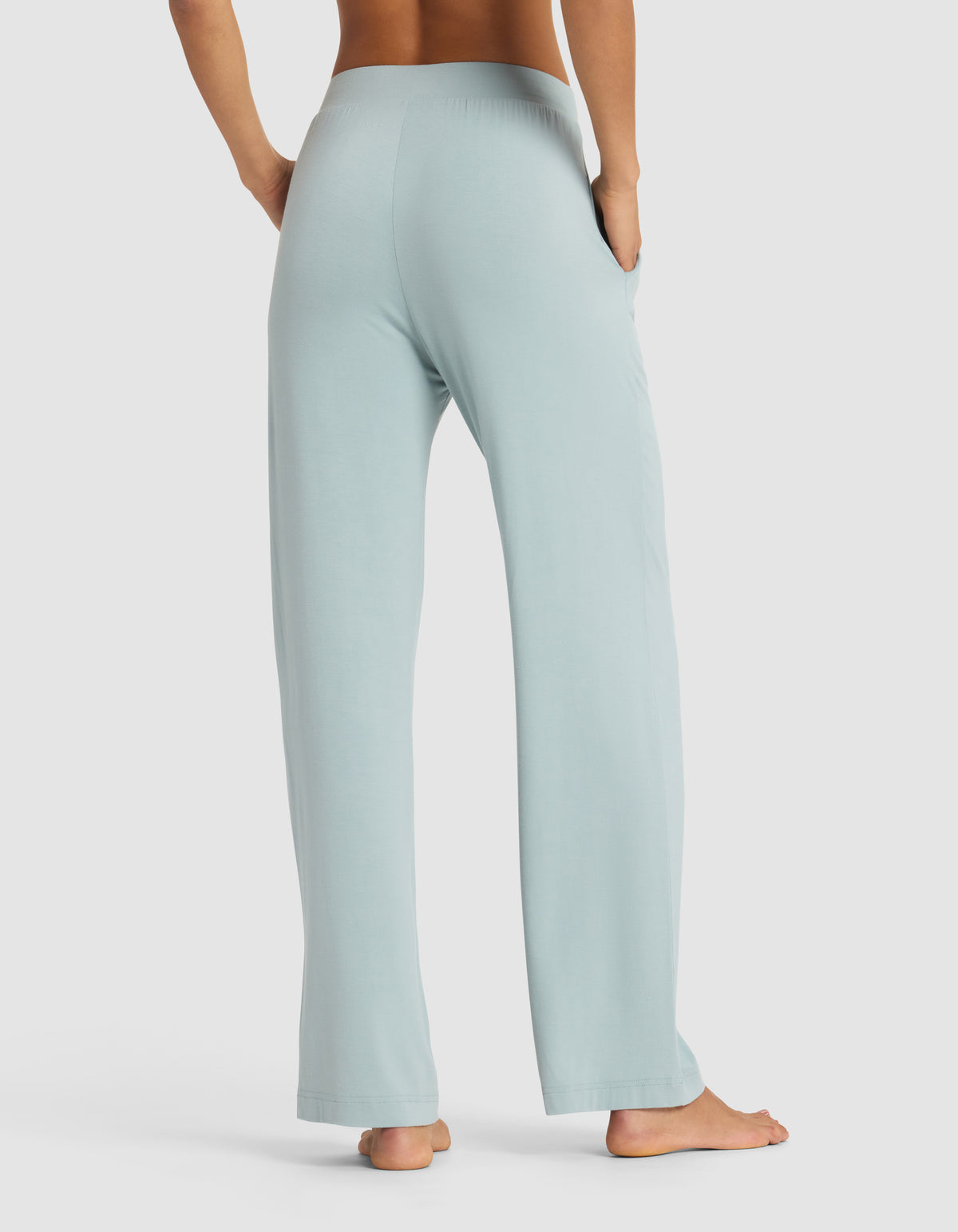 A woman stands barefoot, viewed from behind, wearing Cozy Earth’s Women's Bamboo Stretch-Knit Pant in light blue with hands in the side pockets. The background is plain white.