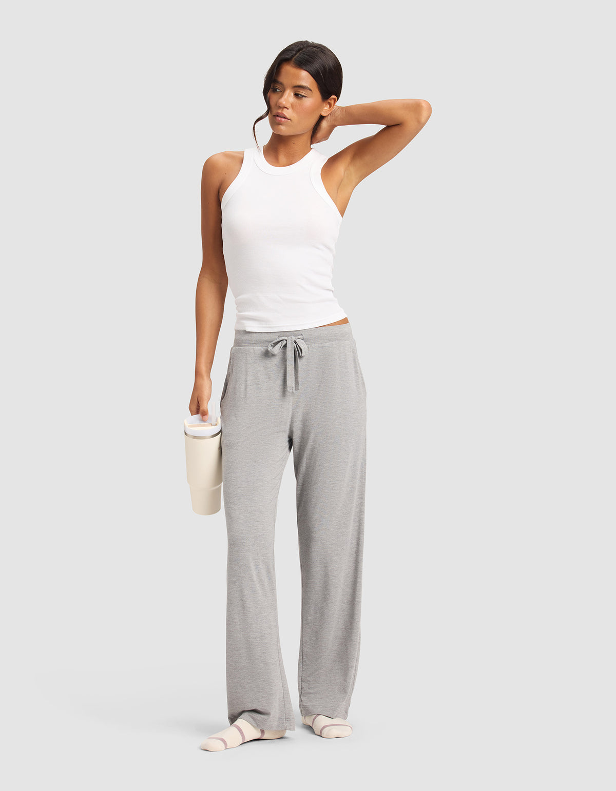 A woman in Cozy Earth's Women's Bamboo Stretch-Knit Pant stands against a plain background, holding a cup with one hand and resting her other hand on her head. She wears a white sleeveless top, gray lounge pants, and light slippers.