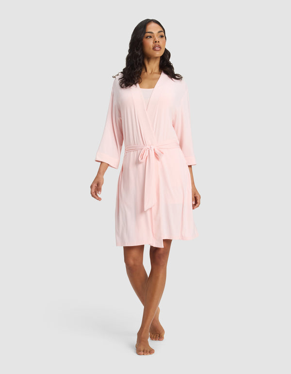 A woman wears the Cozy Earth Women's Bamboo Stretch-Knit Kimono Robe in light pink, knee-length with a belt, standing barefoot against a plain light gray background.|Color:Blossom