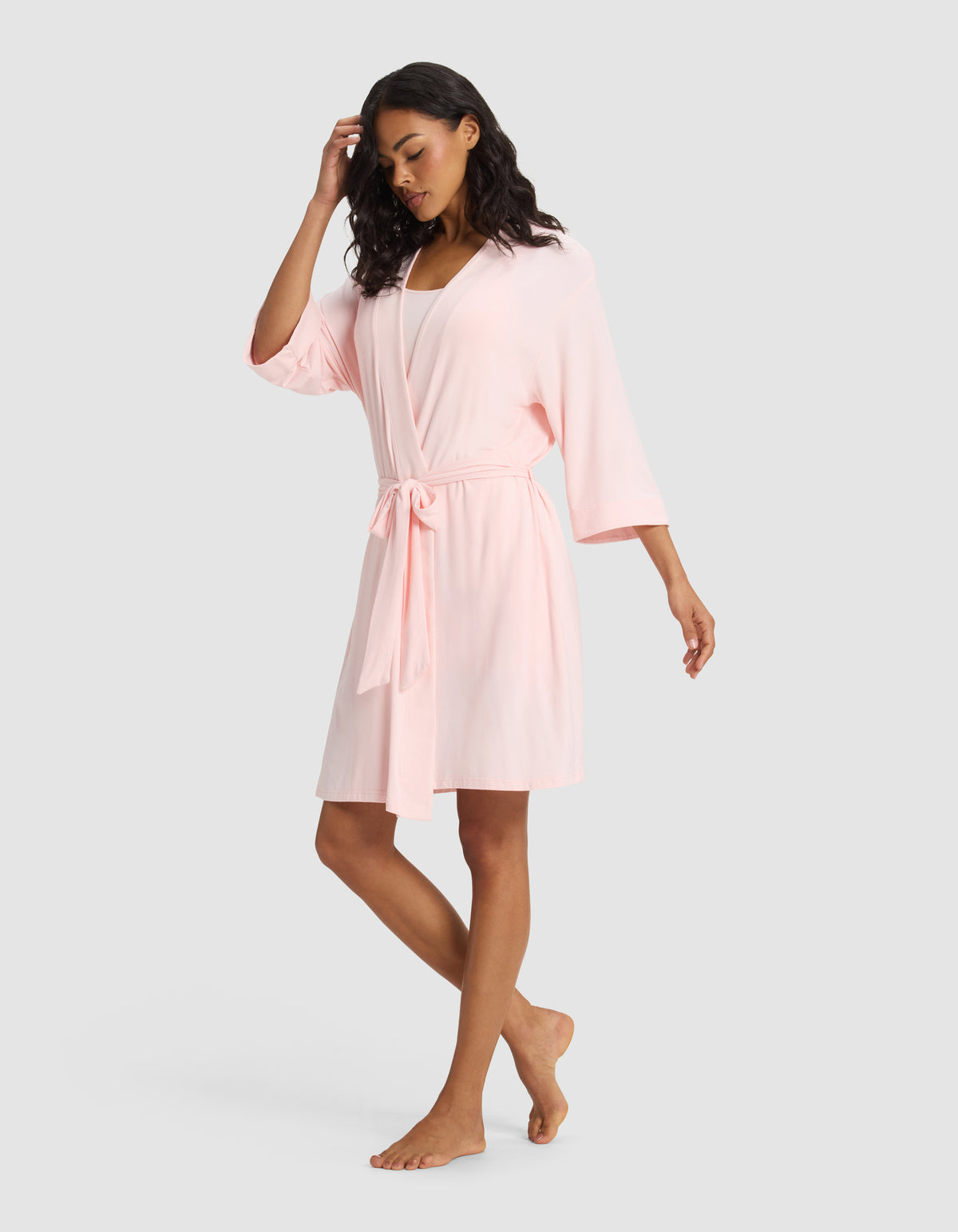 A woman stands barefoot against a plain background, wearing the Cozy Earth Women's Bamboo Stretch-Knit Kimono Robe in light pink, featuring wide sleeves and a waist tie. One hand touches her hair while the other rests by her side.