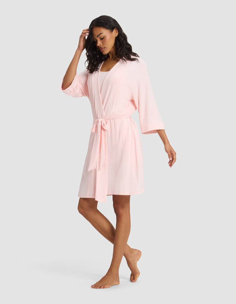 A woman stands barefoot against a plain background, wearing the Cozy Earth Women's Bamboo Stretch-Knit Kimono Robe in light pink, featuring wide sleeves and a waist tie. One hand touches her hair while the other rests by her side.
