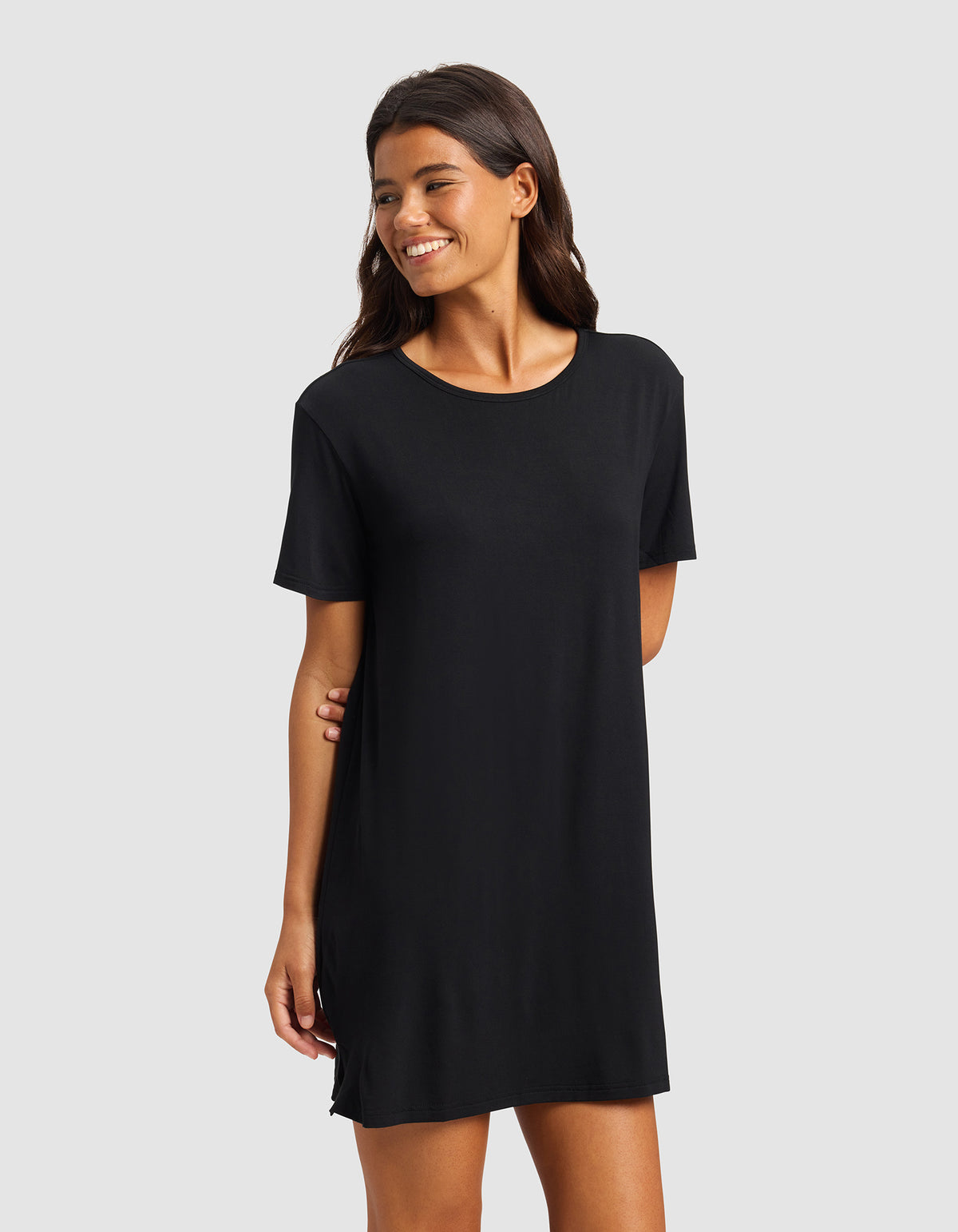 A woman with long dark hair smiles while wearing the Cozy Earth Women's Bamboo Stretch-Knit Sleep Dress, a simple black short-sleeved dress that falls above the knees, standing against a plain light gray background.