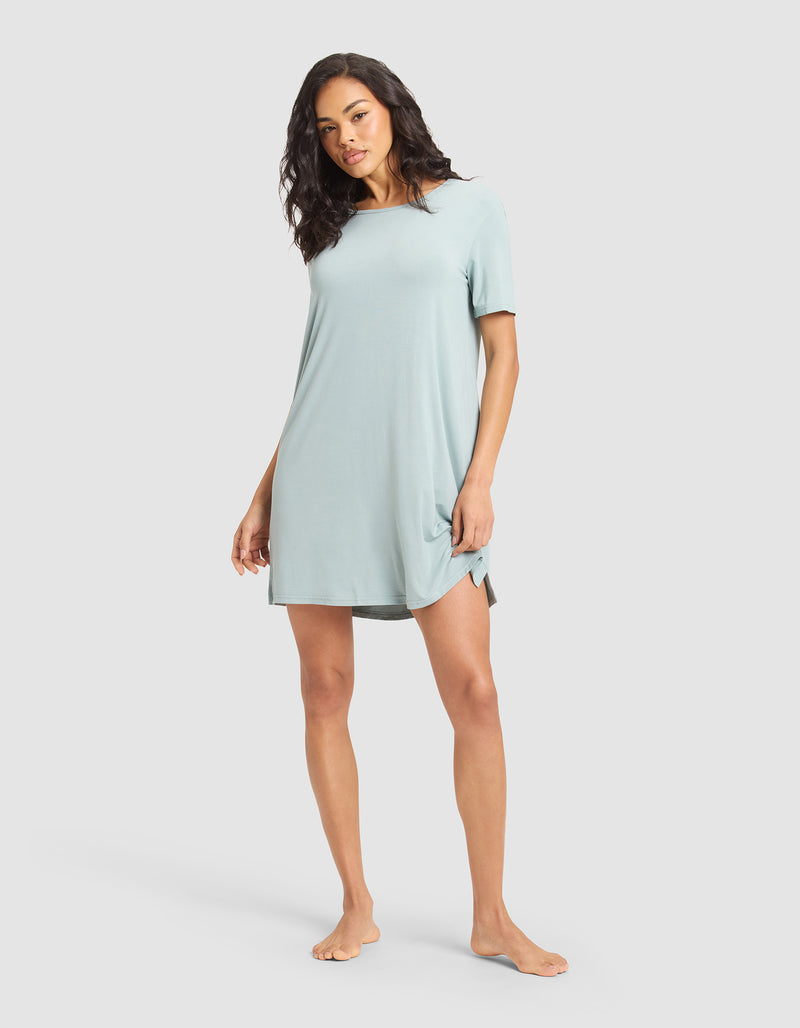 A woman stands barefoot wearing Cozy Earth's Women's Bamboo Stretch-Knit Sleep Dress, a relaxed-fit, light blue short-sleeve dress with a slight side slit, against a plain light gray background.