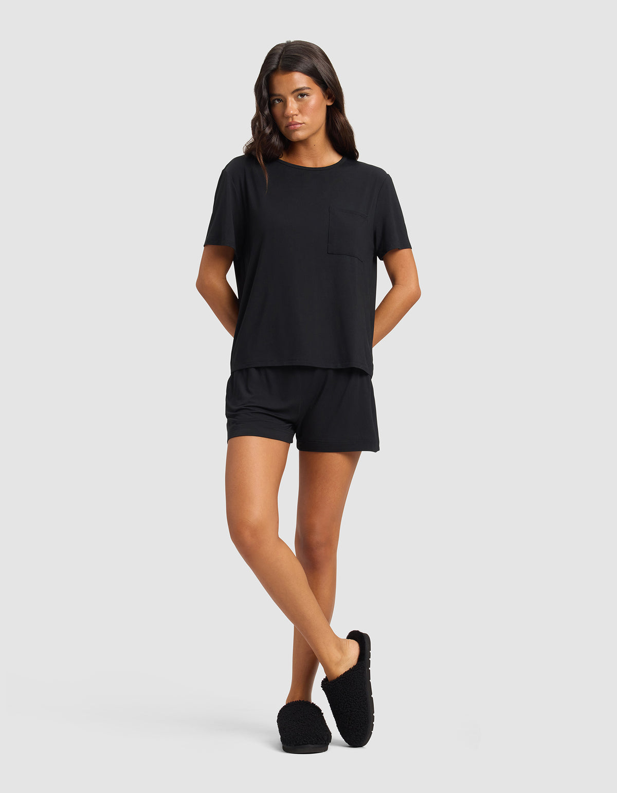 A woman stands against a plain background wearing the Cozy Earth Women's Bamboo Stretch-Knit Lounge Tee in black, paired with matching shorts and slippers. Her arms are relaxed behind her back as she gazes forward.