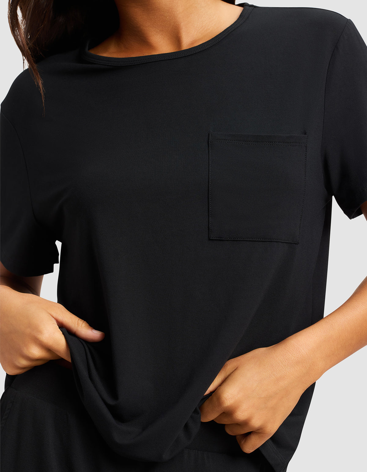 A woman is shown from shoulders to waist wearing the Cozy Earth Women's Bamboo Stretch-Knit Lounge Tee, a plain black short-sleeve shirt with a chest pocket, gently pulling the hem with both hands.