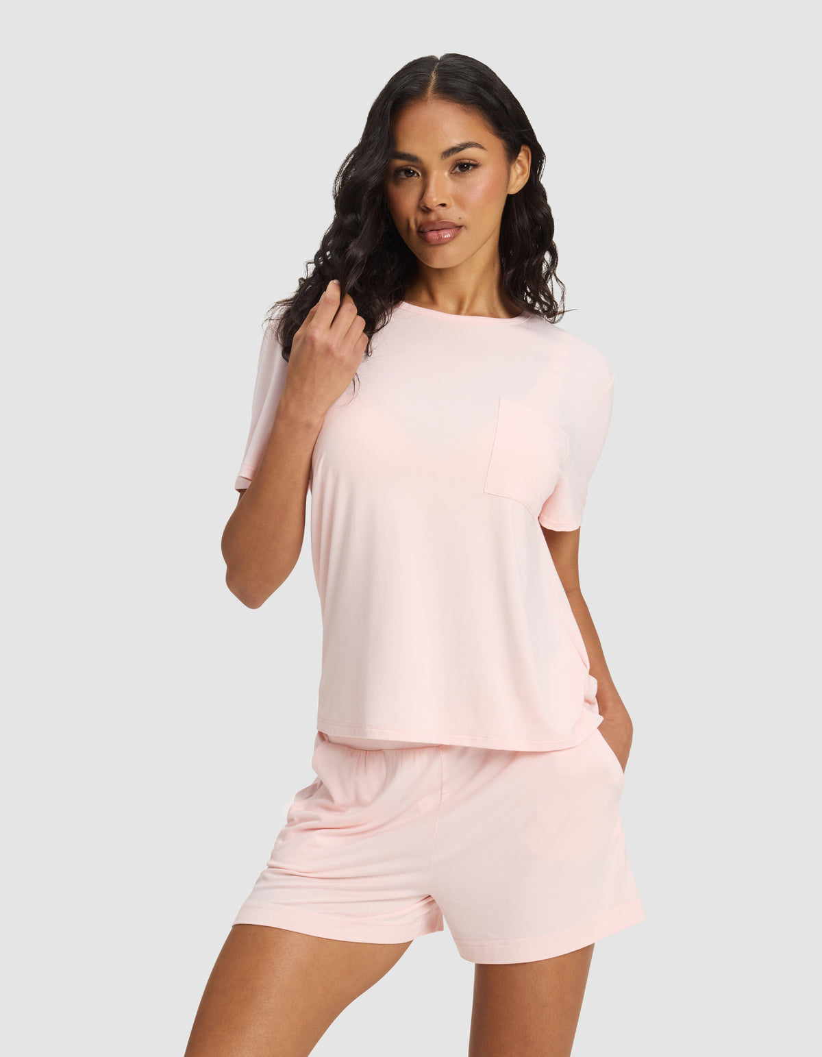 A woman with long, wavy dark hair and a relaxed, confident look stands against a plain background wearing Cozy Earth's Women's Bamboo Stretch-Knit Lounge Tee in light pink with a pocket, paired with matching pink shorts.