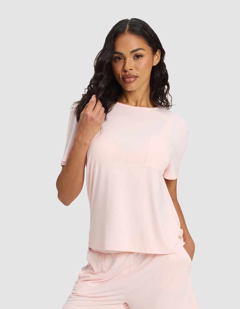 A woman with long wavy hair wears the Cozy Earth Women's Bamboo Stretch-Knit Lounge Tee & Pant Set, light pink in color, one hand in her pocket and the other touching her hair against a plain background.