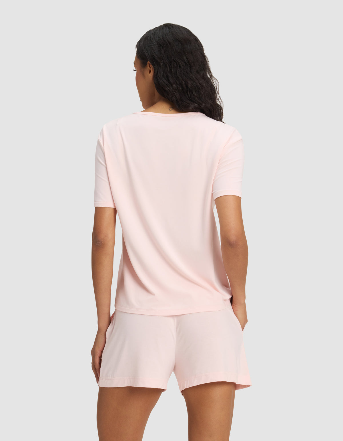 A woman with wavy dark hair is seen from the back, modeling the Cozy Earth Women's Bamboo Stretch-Knit Lounge Tee in light pink, paired with matching shorts against a plain light gray background.