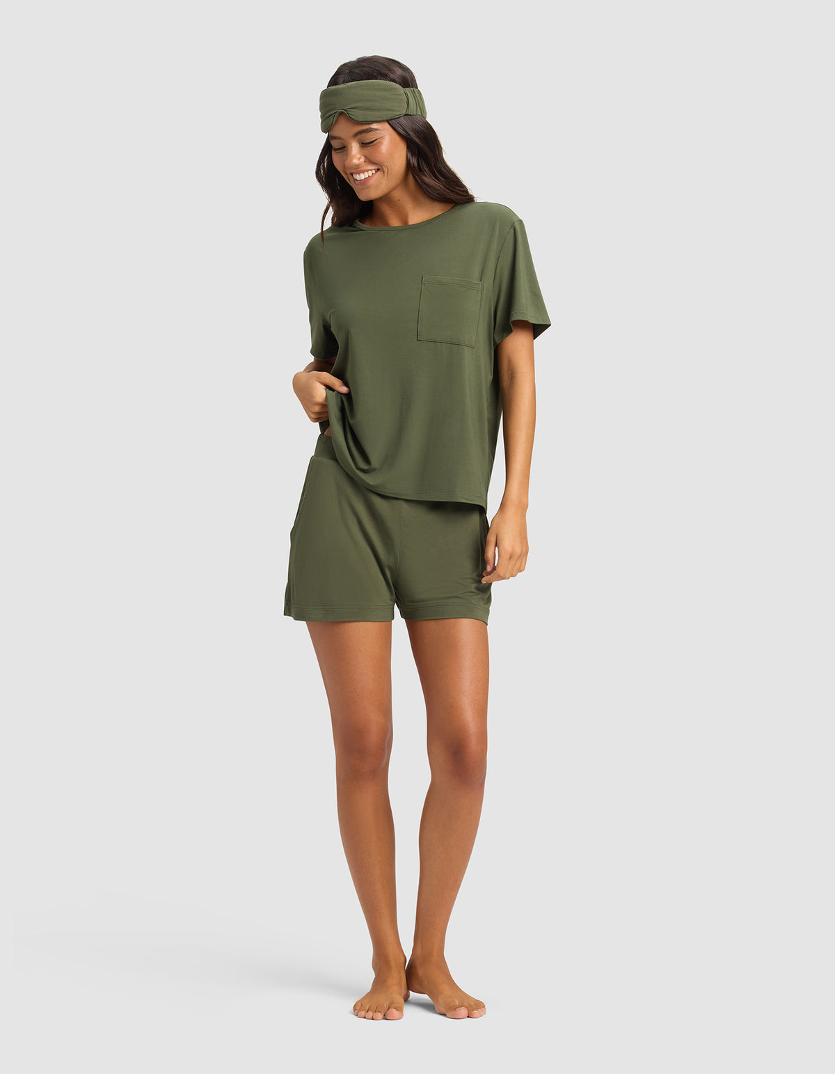 A person stands barefoot against a light gray background, smiling with one hand in their pocket, wearing the Cozy Earth Women's Bamboo Stretch-Knit Lounge Tee in olive green with matching shorts and a headband.