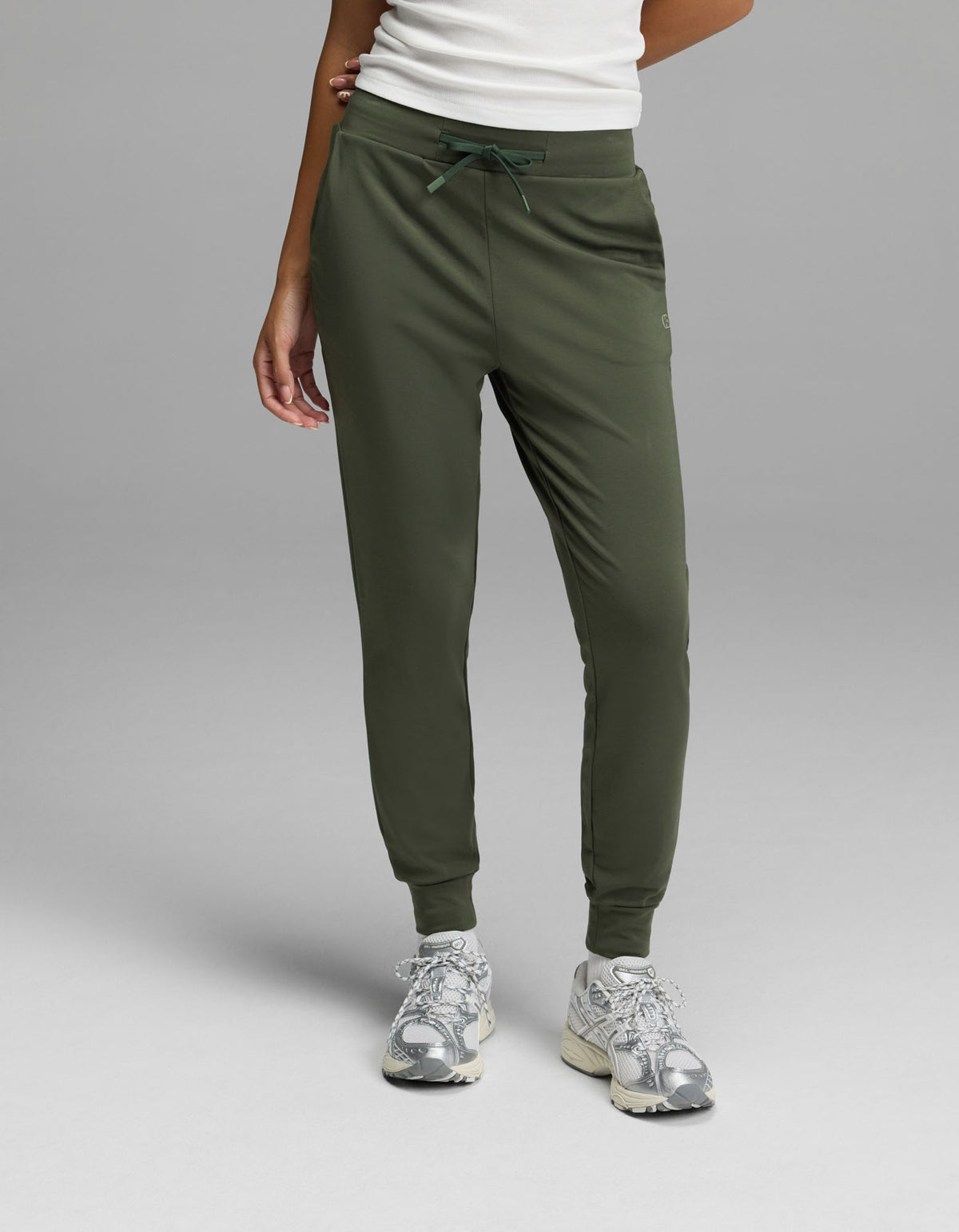 A person is shown from the waist down, wearing Cozy Earth’s Women’s Brushed Bamboo Jogger in olive green with a white top and silver sneakers, standing against a plain gray background. Only the lower body and part of one arm are visible.