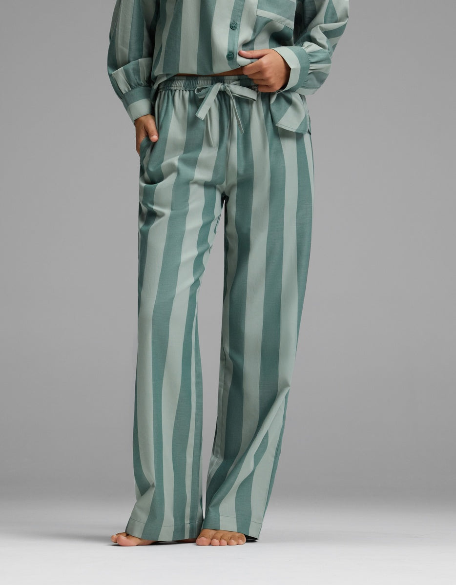 A woman is seen standing barefoot against a plain gray background, wearing Cozy Earth’s Women’s Belmont Pajama Pant in green with vertical stripes, paired with the matching long-sleeve top.|Color:Rosemary Stripe