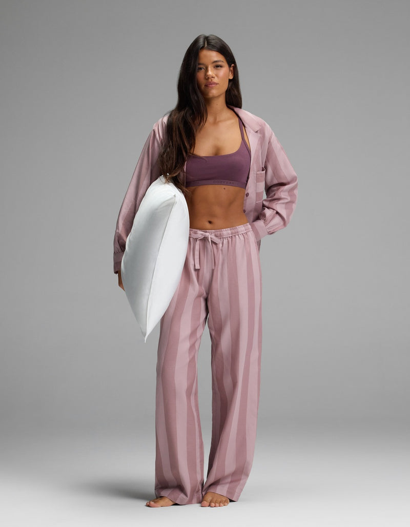 A woman wearing Cozy Earth’s Women’s Belmont Pajama Pant and a purple sports bra stands barefoot on a gray background, holding a white pillow under her arm and looking directly at the camera.