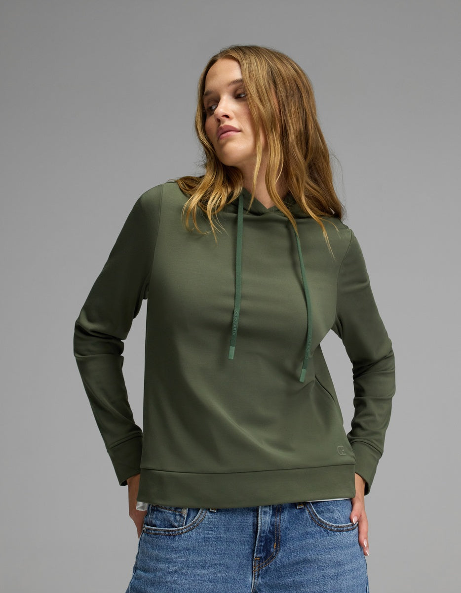 A woman wearing the Cozy Earth Women’s Brushed Bamboo Hoodie in green, paired with blue jeans, stands against a plain gray background, gazing slightly to the side with a relaxed expression.|Color:Olive