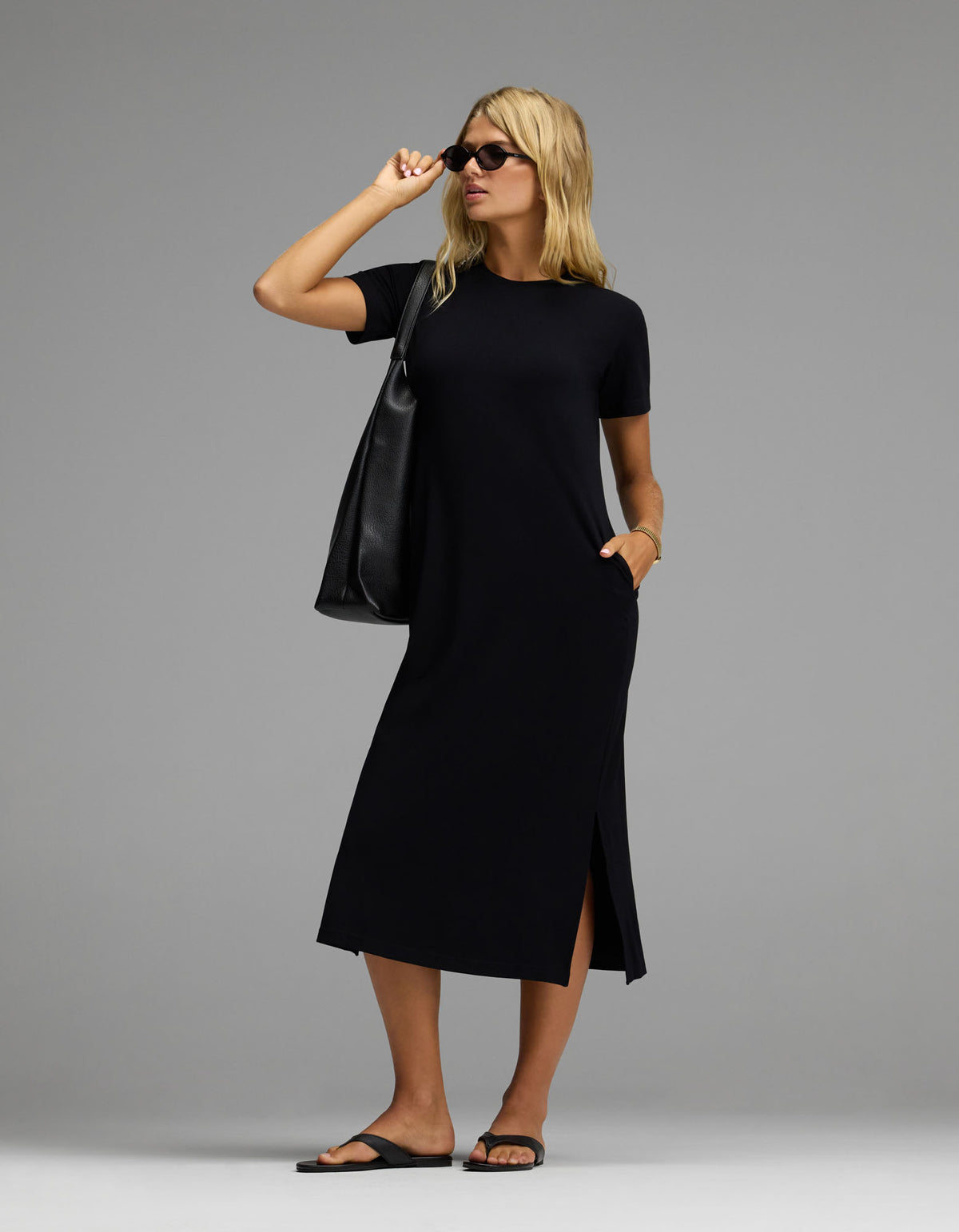 A woman stands confidently against a plain gray background, wearing the Cozy Earth Women’s Brushed Bamboo Midi Dress in black with short sleeves and side slits. She completes her look with black sandals, sunglasses, and a large black tote bag.