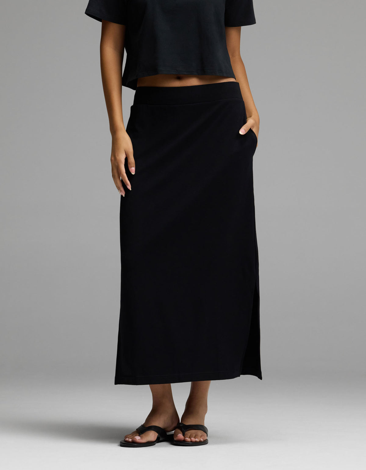A woman wearing Cozy Earth’s Women’s Brushed Bamboo Midi Skirt in black, paired with a black crop top and black sandals, stands with one hand in her skirt pocket against a plain gray background.