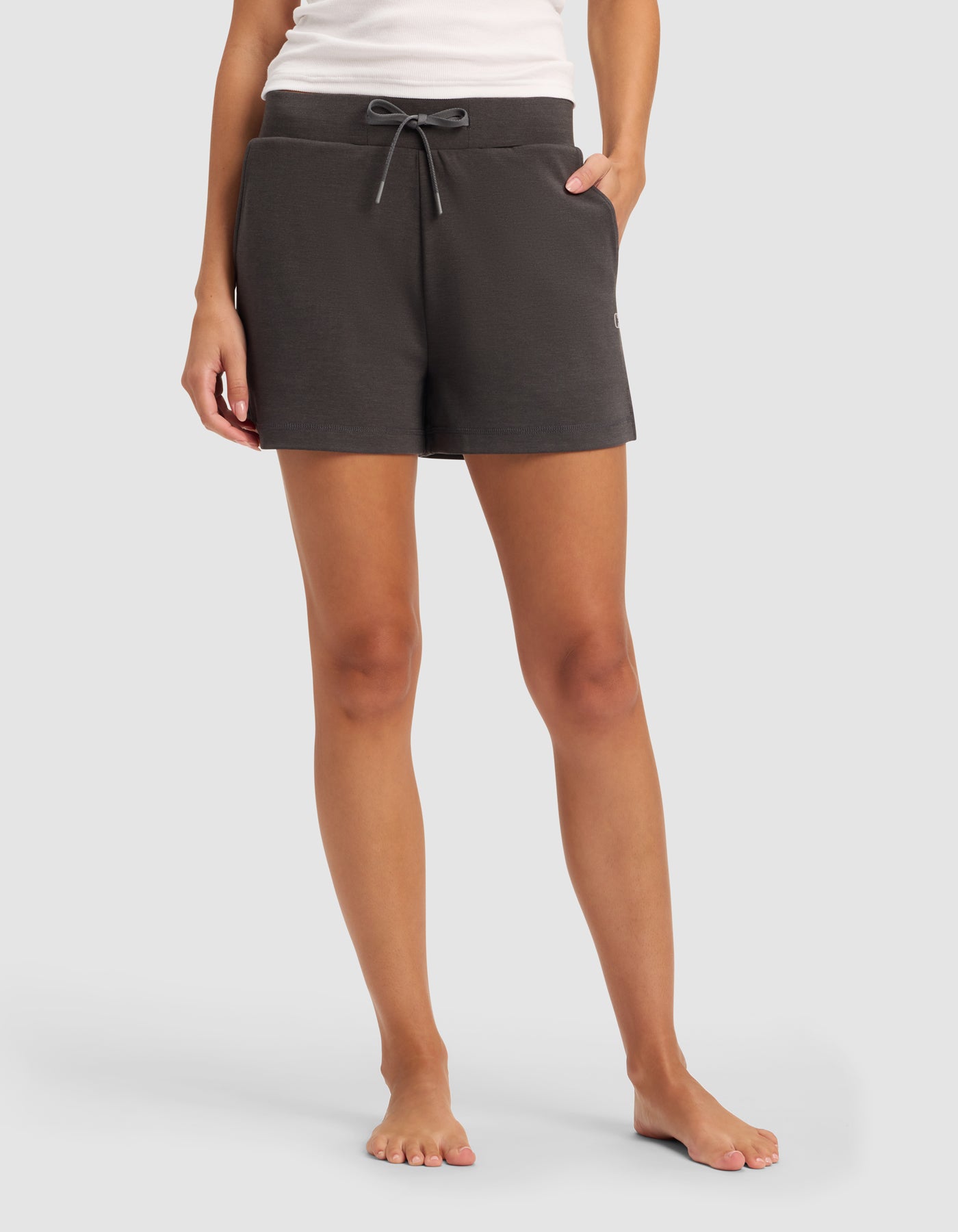 A woman stands barefoot against a plain light gray background, wearing Cozy Earth’s Women’s Brushed Bamboo Shorts in dark gray along with a white tank top.|Color:Charcoal
