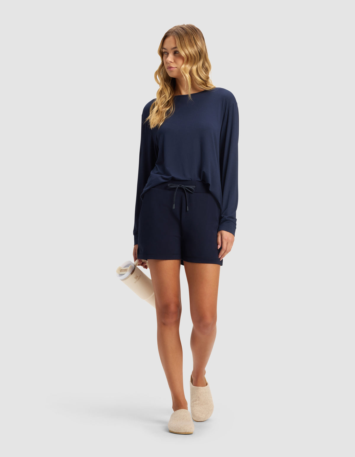 A woman with long wavy hair wears Cozy Earth’s Women’s Brushed Bamboo Shorts in navy blue with a matching long-sleeve top, holds a rolled-up towel, and dons beige slippers against a plain light gray background.