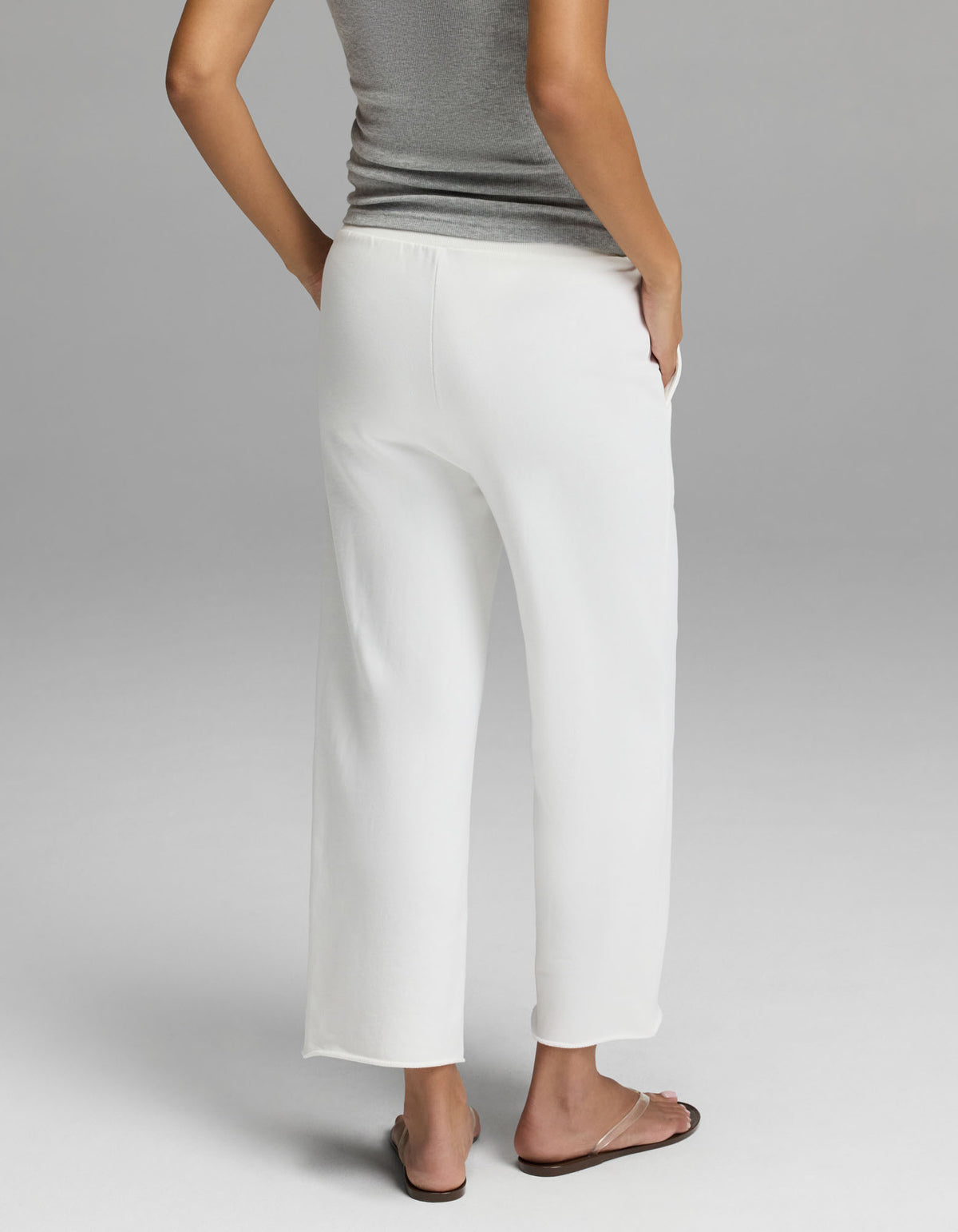A woman is photographed from behind against a plain light gray background, standing with one hand in the pocket of her loose white Cozy Earth Women’s Chelsea Cropped Pant, paired with a gray sleeveless top and flip-flops.|Color:Bone