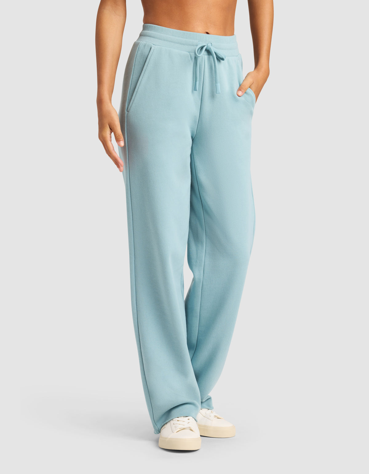 A person models the Cozy Earth Chelsea Pant in light blue with pockets and drawstring, paired with off-white sneakers, standing against a plain light background. The upper body is partially out of frame.
