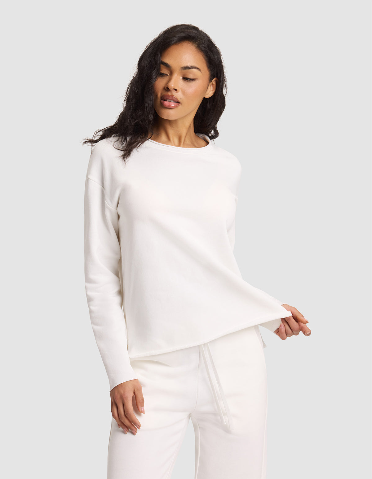 A woman poses against a light gray background wearing the Cozy Earth Women's Chelsea Pullover and matching pants in all white, holding the hem of the top with one hand while looking slightly downward. 