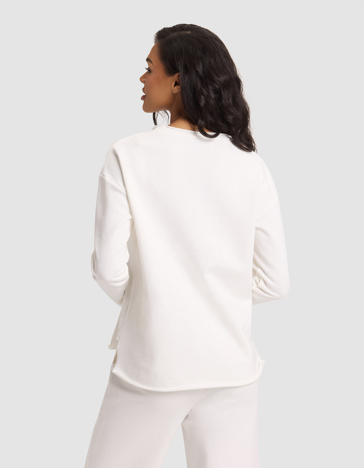 A woman with medium-length dark hair is seen from behind wearing Cozy Earth's Women's Chelsea Pullover in white, paired with white pants, standing against a plain light gray background. |Color:Bone