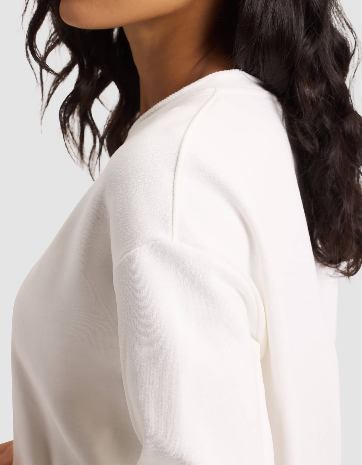 A woman with wavy dark hair wears the Cozy Earth Women's Chelsea Pullover, an off-white long-sleeve top, shown from the shoulders up and slightly turned to the side against a plain light background. 