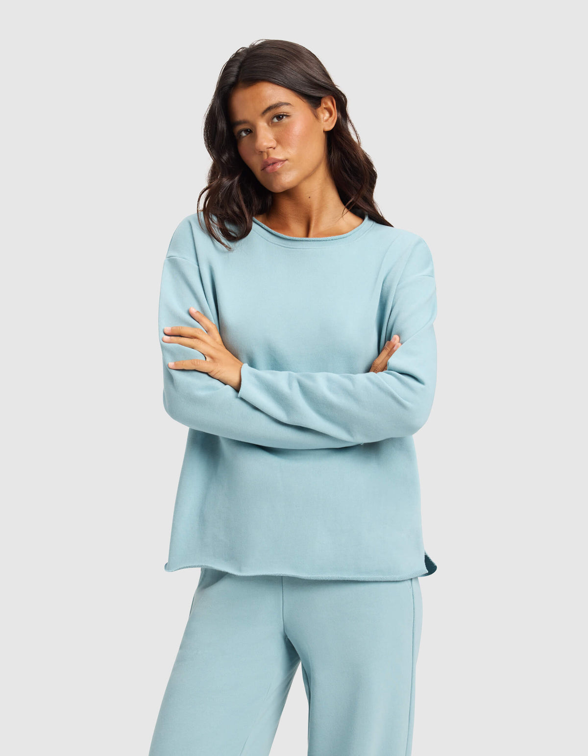 A woman with long dark hair stands with arms crossed, wearing the Cozy Earth Women's Chelsea Pullover and matching pants in light blue against a plain light gray background.