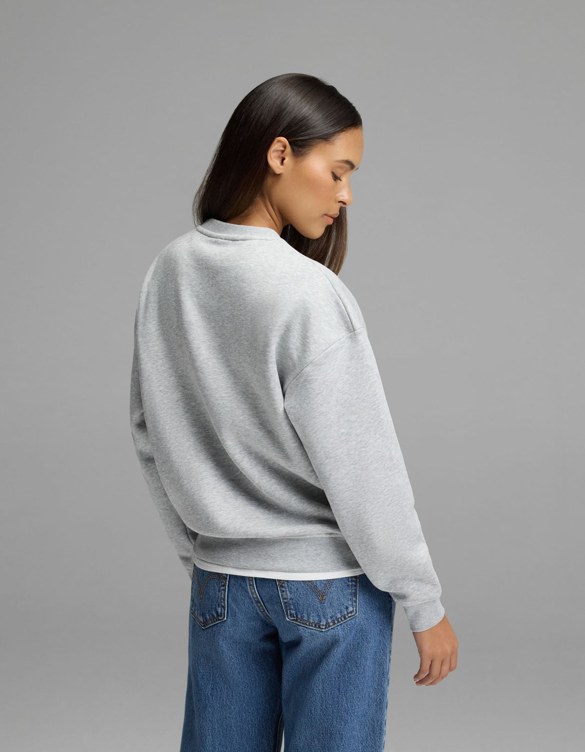 A woman with long dark hair, wearing the Cozy Earth Women’s Cityscape Crewneck in light gray and blue jeans, stands facing away from the camera against a plain gray background.