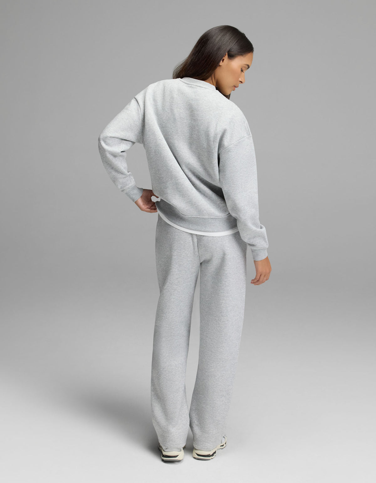 A person stands facing away, dressed in the Cozy Earth Women’s Cityscape Crewneck & Wide Leg Pant Set in light gray with white sneakers, against a plain gray background.