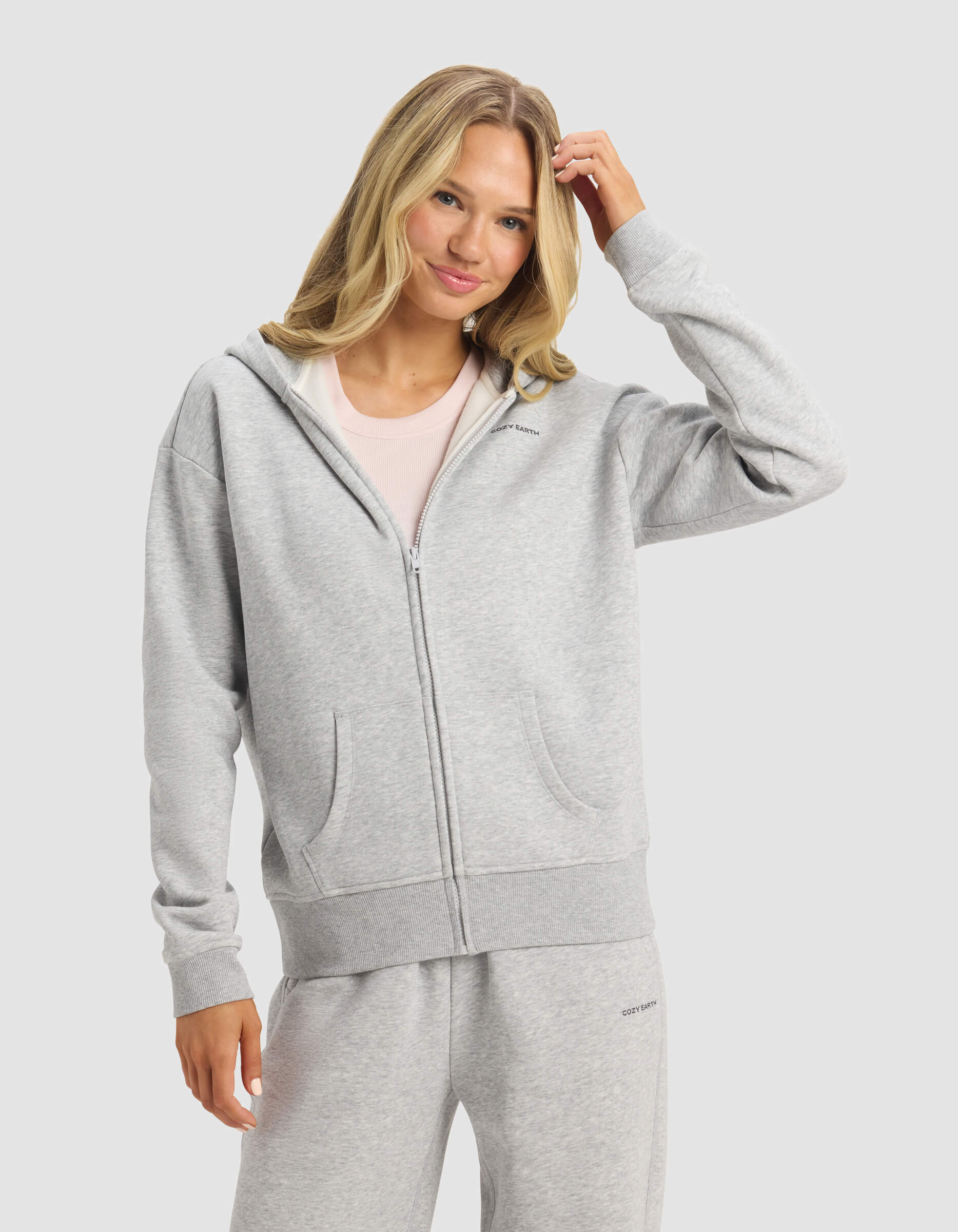 A woman with long blonde hair smiles and touches her hair while wearing Cozy Earth's Women's CityScape Full Zip hoodie and matching sweatpants, standing against a plain light background.|Color:Heather Grey