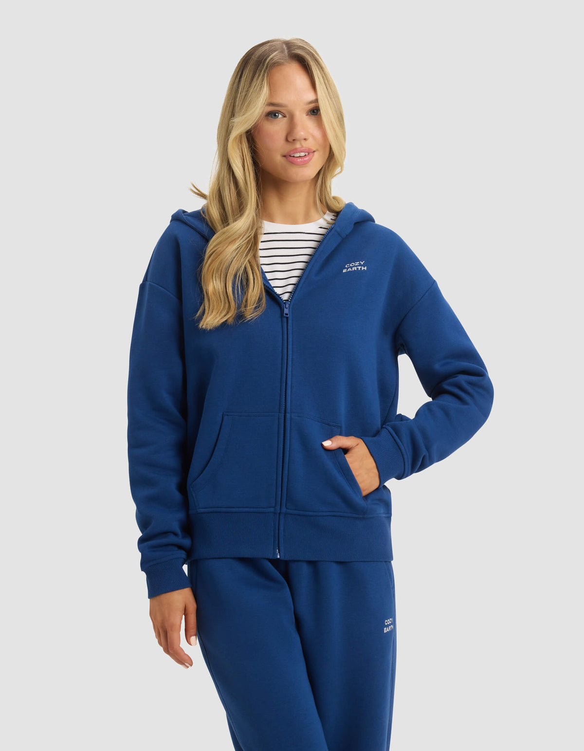A woman with long blonde hair models the Cozy Earth Women's CityScape Full Zip hoodie and matching blue sweatpants over a white and navy striped shirt, posing with one hand in her pocket against a plain light background.