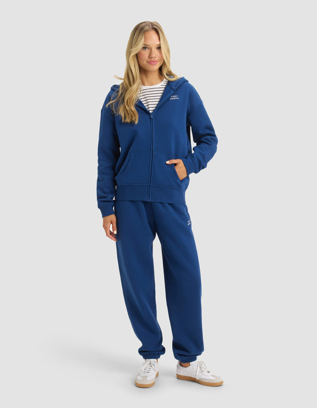 A woman with long blonde hair smiles slightly against a plain background, wearing Cozy Earth's Women's CityScape Full Zip hoodie in blue, matching sweatpants, a striped shirt underneath, and white sneakers.