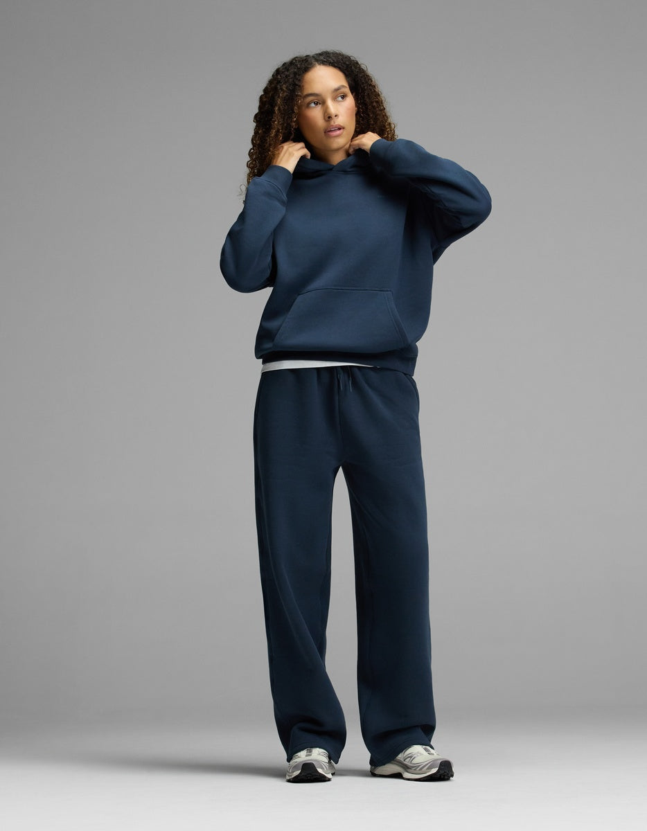 A person with curly hair adjusts the hood of the Women’s CityScape Hoodie & Wide Leg Pant Set by Cozy Earth in navy blue, standing against a plain gray background.|Color:Eclipse