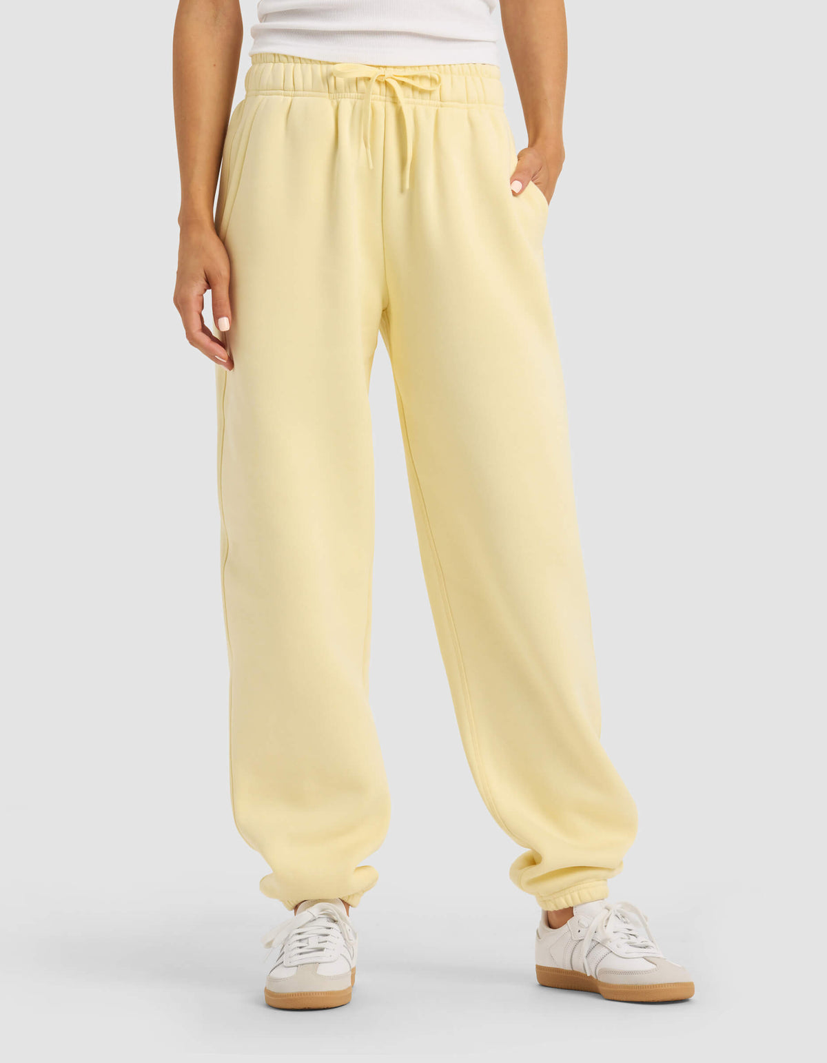 A person stands against a plain background with hands in the pockets of Cozy Earth Women's Cityscape Sweatpant in light yellow, paired with a white top and white sneakers.