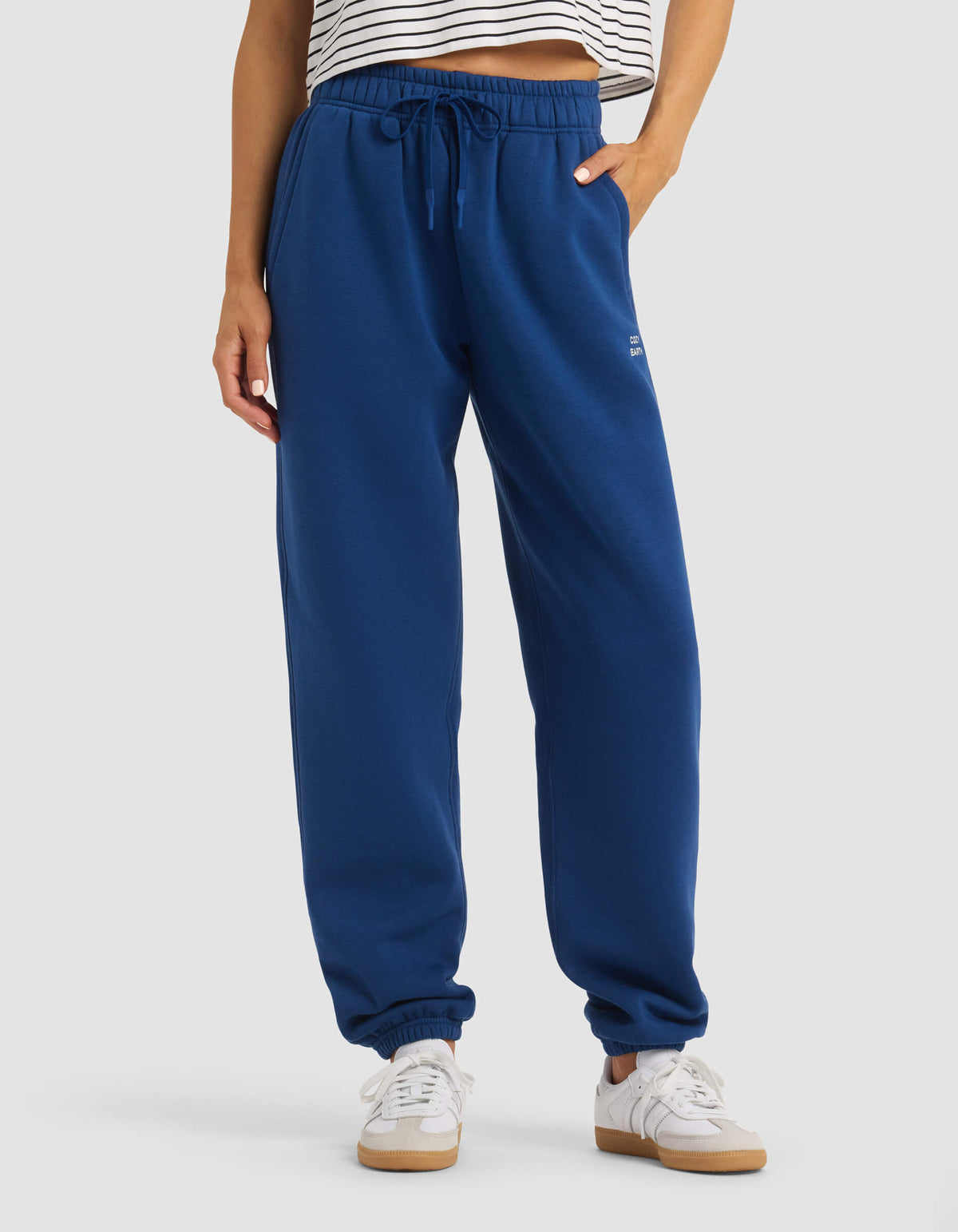 A person wearing Cozy Earth's Women's Cityscape Sweatpant in royal blue, paired with a striped crop top and white sneakers, stands against a plain background, showing only their lower torso and legs.