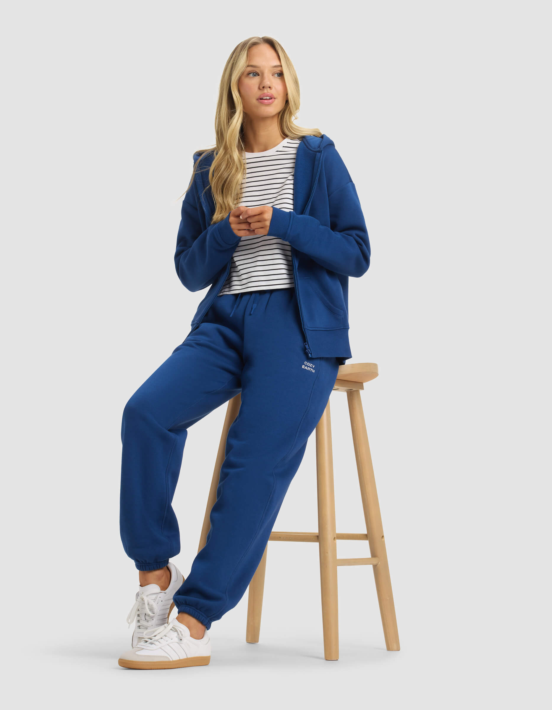 A woman with long blonde hair wears Cozy Earth's Women's Cityscape Sweatpant in blue, paired with a white and black striped shirt and white sneakers. She sits on a wooden stool, hands together, looking slightly to the side.|Color:Pacific