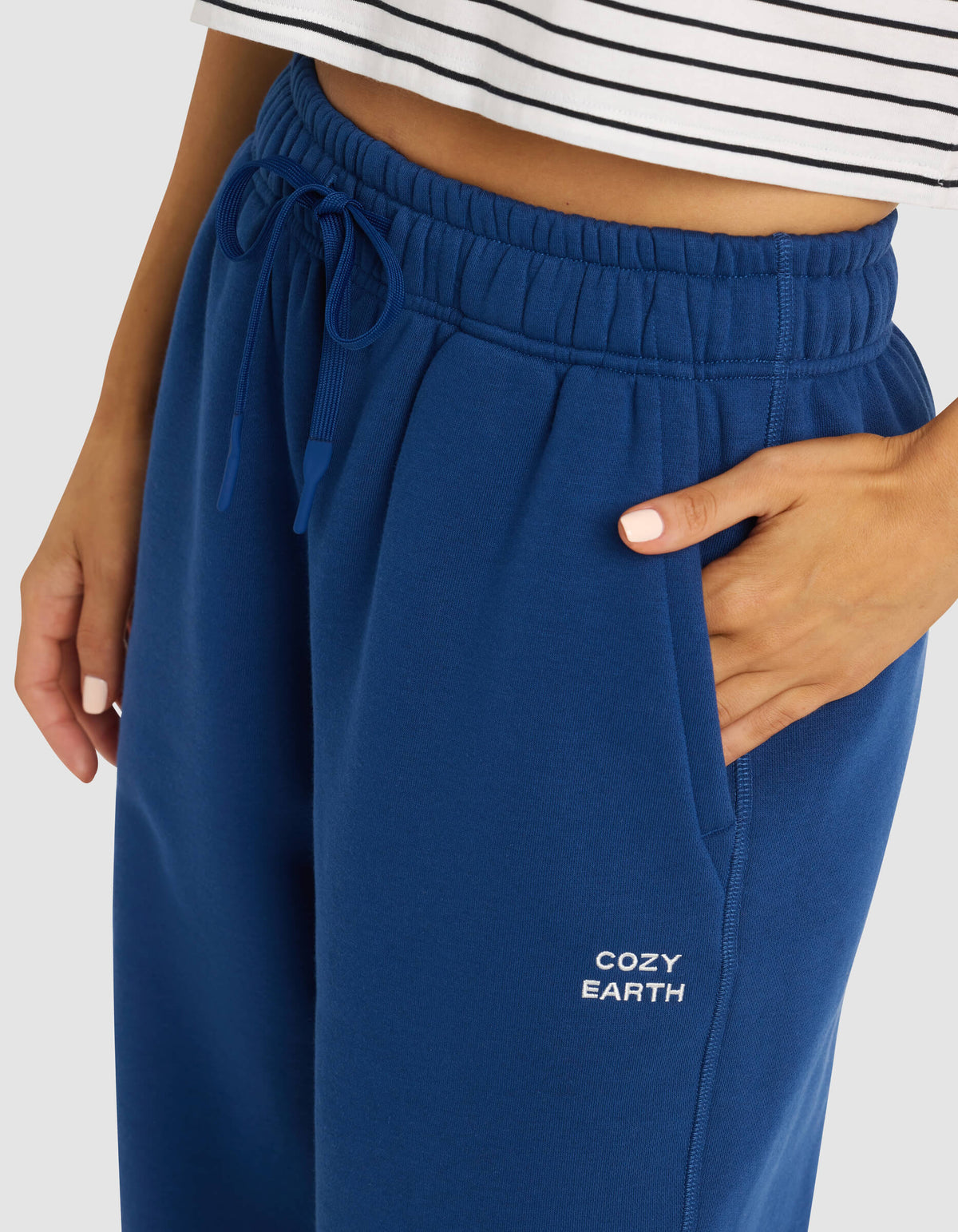 A woman wears Cozy Earth's Women's Cityscape Sweatpant in blue, featuring the brand name on the pocket, paired with a white striped crop top and one hand in her pocket.