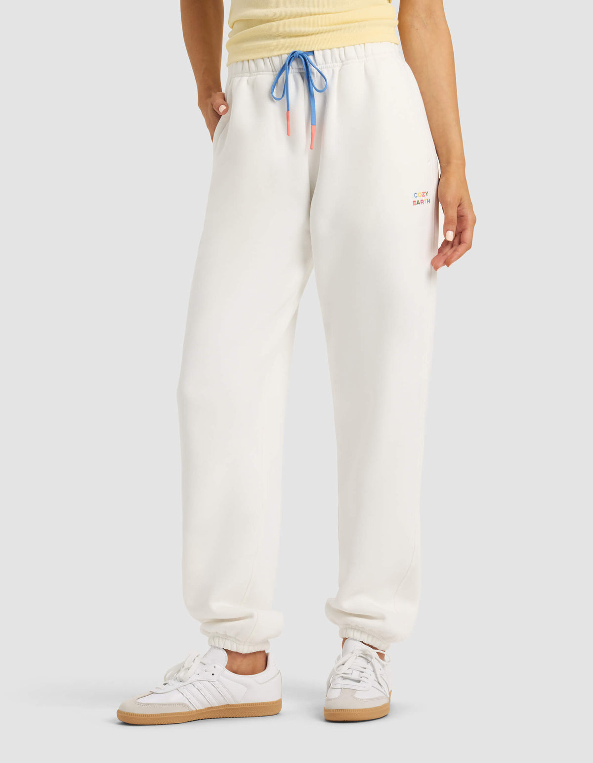A person models Cozy Earth's Women's Cityscape Sweatpant in white, paired with a yellow top and white sneakers, hands in pockets, against a plain background. 