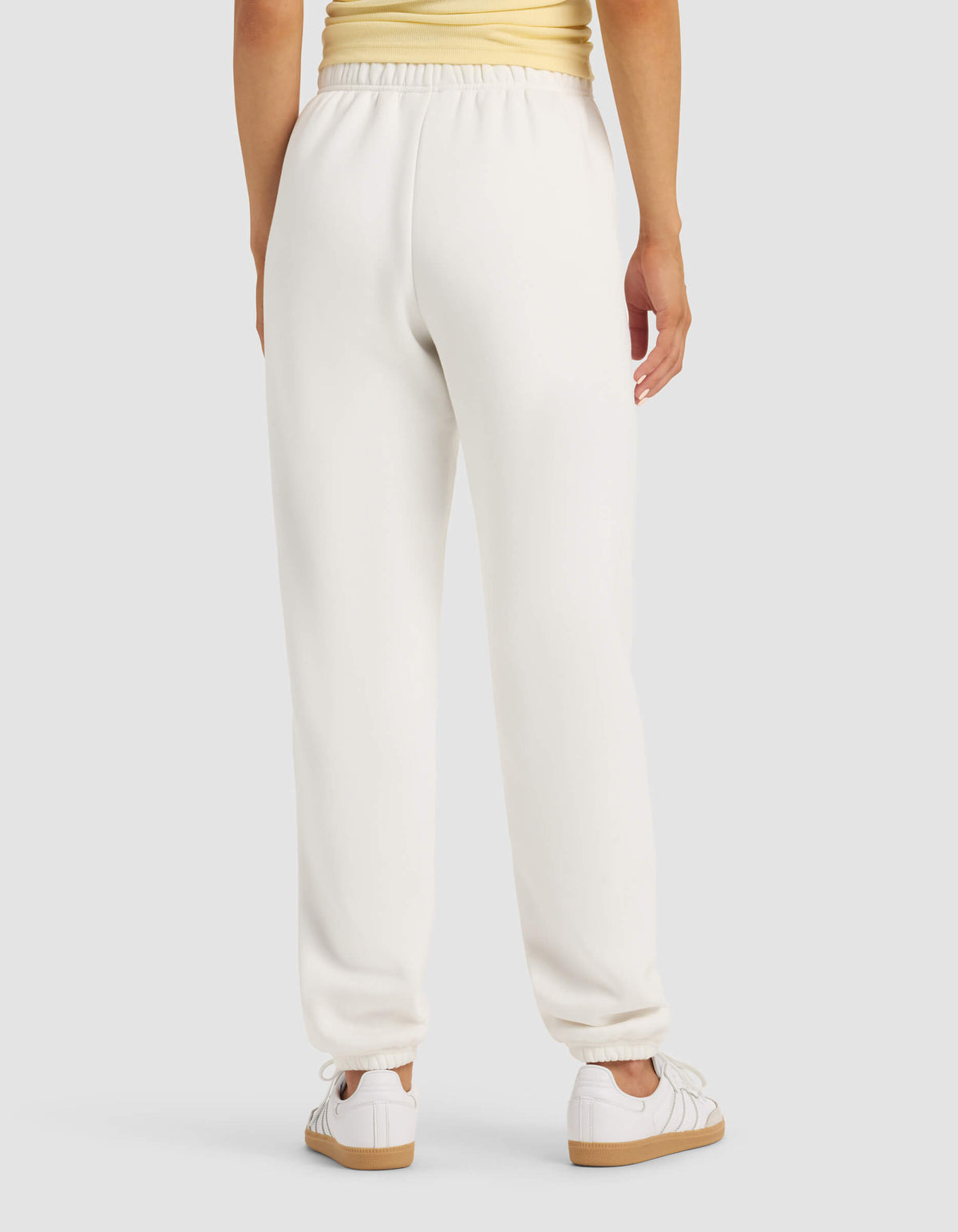 A person is seen from behind wearing the Cozy Earth Women's Cityscape Sweatpant in white, paired with a light yellow top and white sneakers, standing against a plain light gray background.