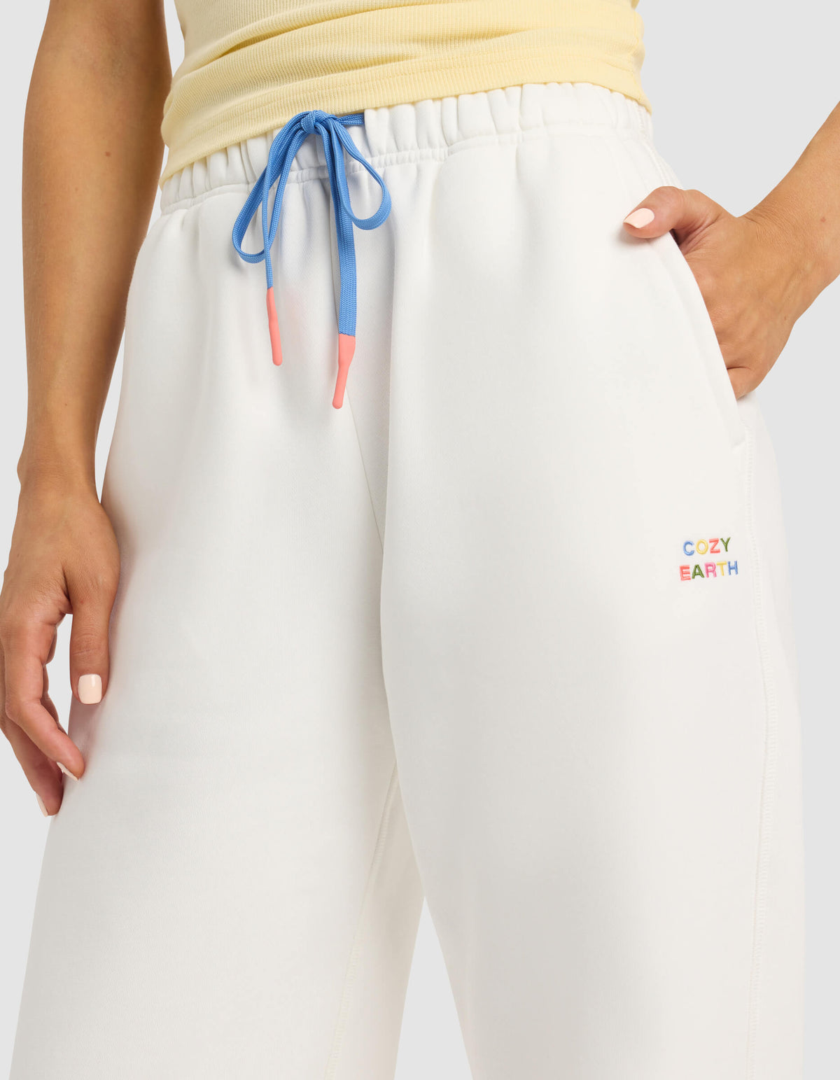 A person wears Cozy Earth Women's Cityscape Sweatpant in white with blue drawstrings and a multicolored logo, standing with one hand in their pocket. The top of a light yellow shirt is also visible.