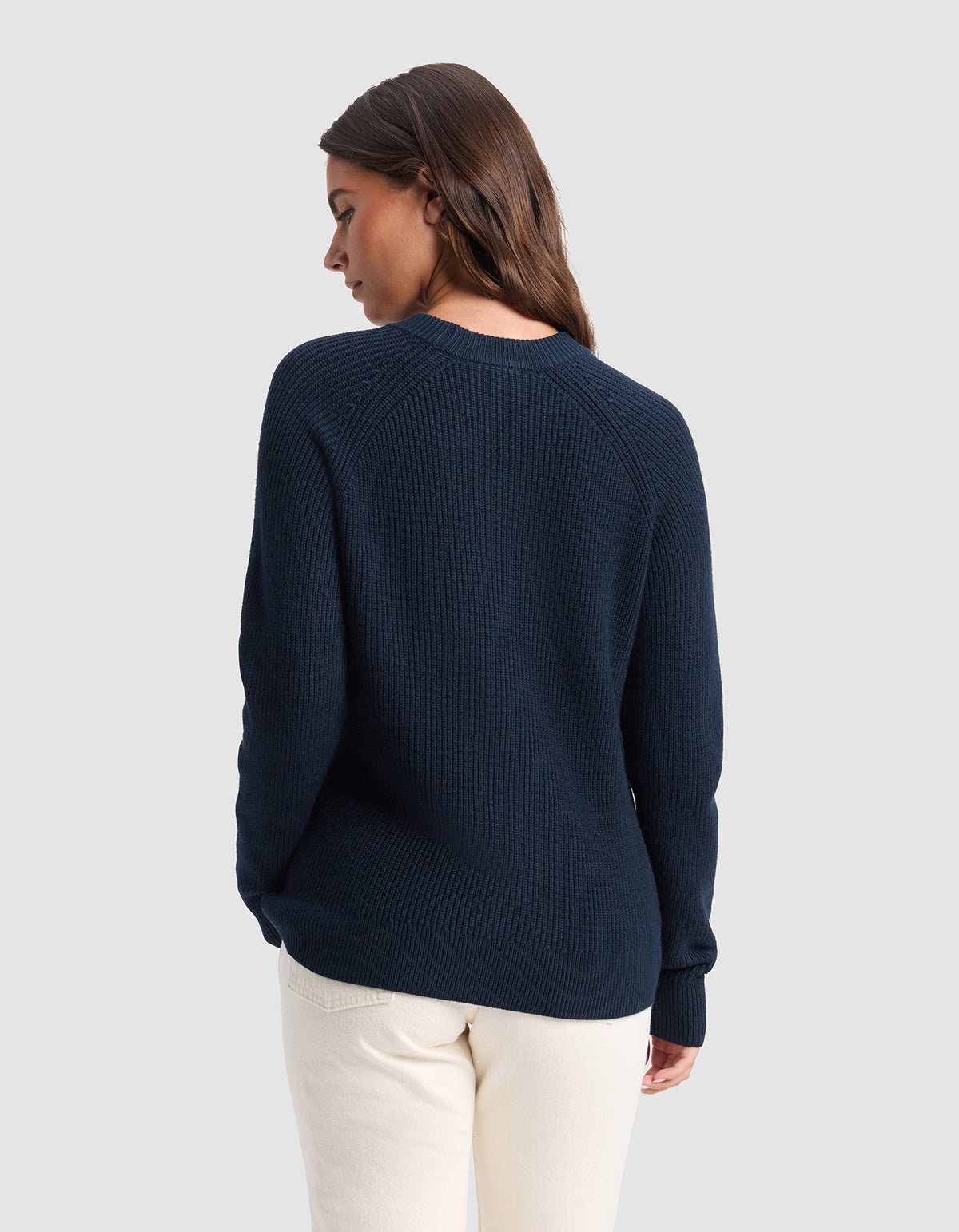 A woman with long brown hair stands with her back to the camera, wearing Cozy Earth's Women's Classic Crewneck in navy blue and off-white pants against a light grey background.