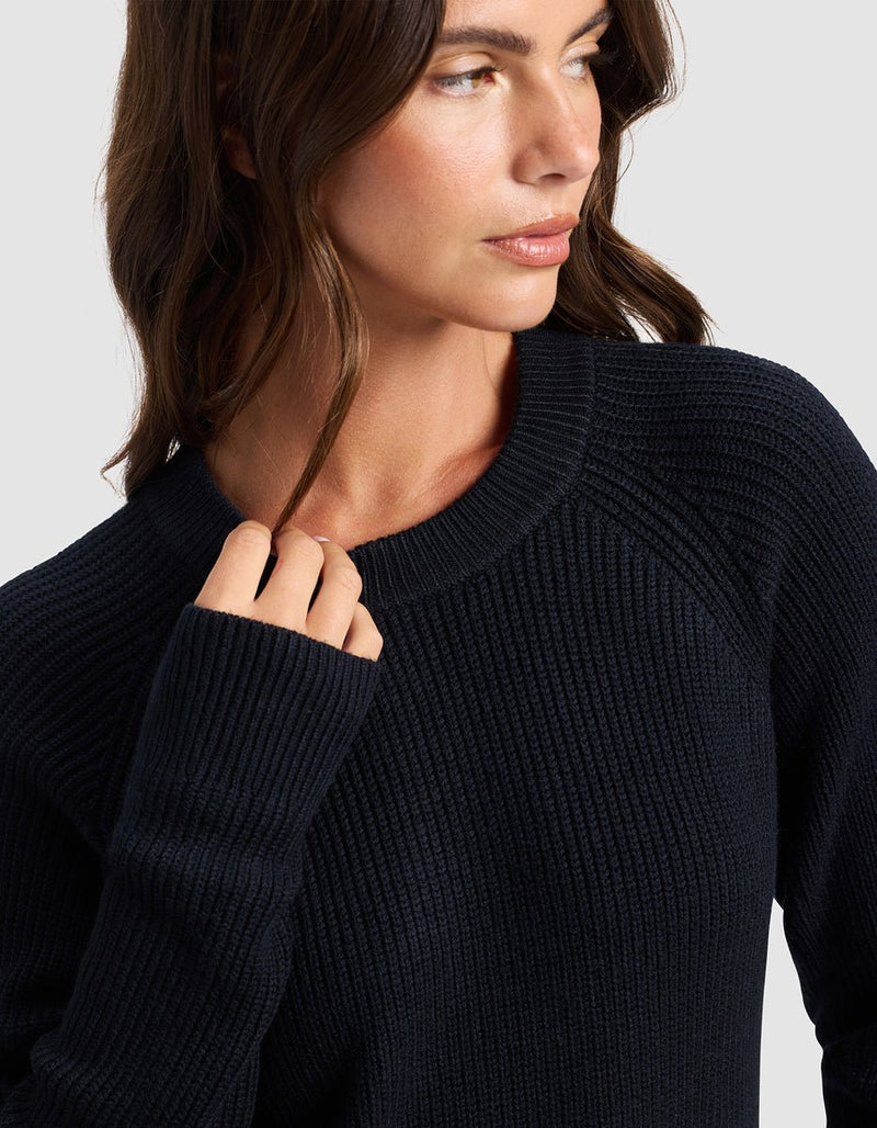 A woman with wavy brown hair wears the Cozy Earth Women's Classic Crewneck in dark navy, facing slightly to the side with her hand near her collar. The background is light gray.