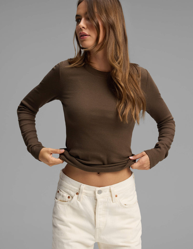 A woman with long brown hair wears the Cozy Earth Women's Fine Ribbed Long Sleeve Tee in brown, paired with high-waisted white jeans, standing against a plain gray background.
